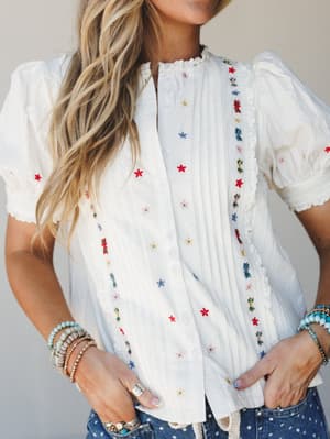 A white embroidered blouse with scattered multicolor floral stitching and lace trim details, styled with stacked beaded bracelets and polka dot jeans — sweet, lived-in, and ready for a sun-filled afternoon.