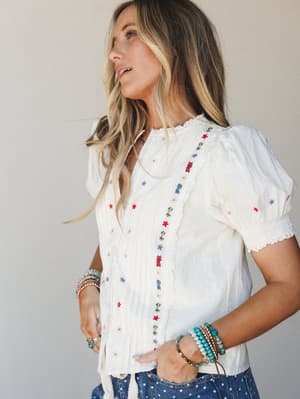 A cream embroidered blouse with scattered floral stitching and lace trim details, styled with denim and stacked turquoise bracelets — the kind of top that makes a Tuesday feel intentional.