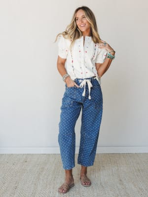 A smiling model pairs a cream embroidered blouse with polka dot cropped cargo jeans and flat sandals — the kind of effortlessly expressive outfit that feels just as good as it looks.