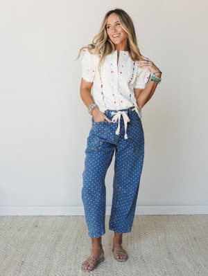 A smiling model pairs a cream embroidered blouse with polka dot cropped cargo jeans and flat sandals — the kind of effortlessly expressive outfit that feels just as good as it looks.