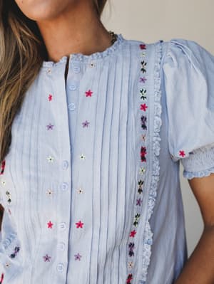 A close-up of a periwinkle blue blouse covered in scattered multicolor floral embroidery, with pintuck pleating down the front, lace trim detailing along the placket, and smocked puff sleeves — the kind of top that makes a simple outfit feel like it