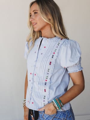 A light blue button-front blouse with scattered floral embroidery and pintuck detailing catches soft natural light — the kind of top that looks equally at home on a patio brunch or a farmers market Saturday.