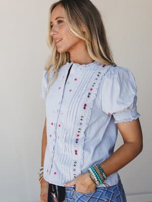 A light blue button-front blouse with scattered floral embroidery and pintuck detailing catches soft natural light — the kind of top that looks equally at home on a patio brunch or a farmers market Saturday.