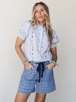 A light and layered warm-weather look — an embroidered misty blue blouse with smocked puff sleeves paired with plaid drawstring bermuda shorts, styled with stacked bracelets and hands tucked in pockets like she's already halfway to somewhere fun.