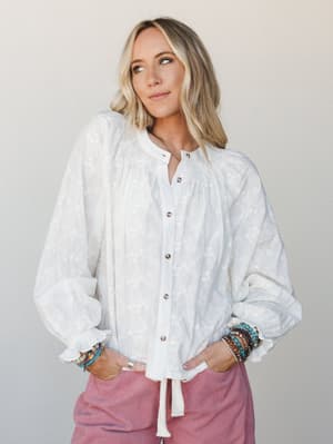 A breezy white floral embroidered blouse with voluminous sleeves and button-front closure, styled with dusty rose corduroy pants and a stack of beaded bracelets — effortlessly soft and lived-in.