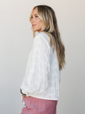A model shot from a three-quarter back angle showing a flowy white-on-white floral embroidered top with billowy sleeves, paired with dusty rose corduroy pants and layered beaded bracelets — soft, effortless, and very much a Sunday morning kind of out