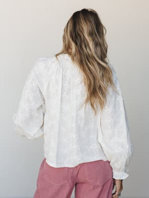 Shot from behind, a model with loose beachy waves wears a cream floral-embroidered blouse with voluminous sleeves paired with dusty rose corduroy pants — soft, romantic, and effortlessly lived-in.