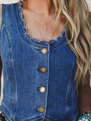 A close-up of a fitted denim vest with scalloped neckline trim and brass buttons, styled with layered gold necklaces and a peek of turquoise beaded bracelets — the kind of piece that makes a simple outfit feel like a whole personality.