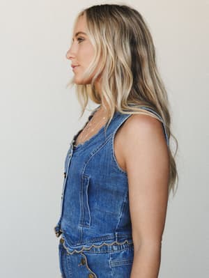 A side-profile studio shot of a model in a medium-wash denim vest top with scalloped hem detail and brass snap buttons — easygoing Western-inspired style that feels intentional without trying too hard.