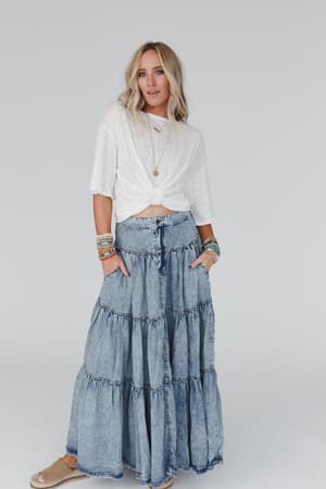 A woman stands against a plain background wearing The Nest Your Favorite Slub Tee Shirt in Natural, paired with a long tiered denim skirt, espadrille shoes, layered necklaces, and colorful bracelets, her hands tucked in her pockets.