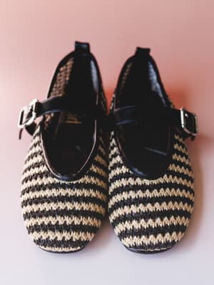 A pair of woven Mary Jane flats in a black and natural stripe pattern, shot overhead against a dusty rose background — the kind of shoes that feel vintage-found but brand new.