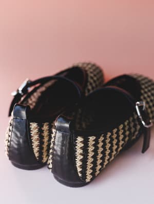 A close-up detail shot of woven flats with black leather trim and silver buckle straps, shot heel-first against a soft dusty rose backdrop — the kind of shoes that feel as good as they look.