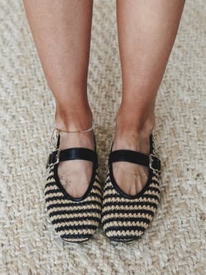 A close-up of woven raffia Mary Jane flats in black and natural stripe, worn on a jute rug — the kind of shoes that make you feel like you planned your whole outfit around them (even if you didn't).
