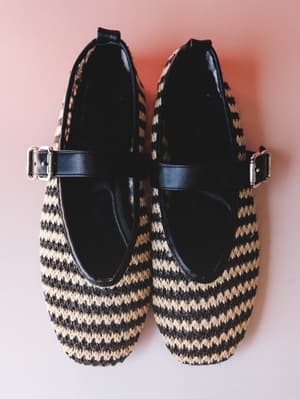 A pair of woven straw Mary Jane flats in black and natural stripe, shot overhead on a dusty pink background — the kind of shoes that make a sundress feel like a whole personality.