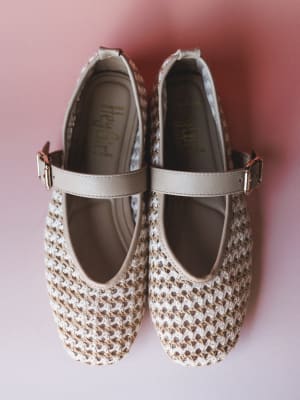 A pair of woven raffia-style Mary Jane flats in natural cream and tan, shot overhead against a soft pink background — the kind of shoes that feel like a warm-weather secret.