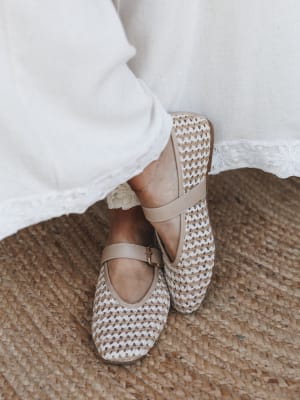 A close-up of woven Mary Jane flats in natural tan and cream, paired with a white dress with floral eyelet hem trim, standing on a jute rug — the whole thing feels like a slow Sunday morning with good decisions.