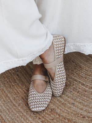 A close-up of woven Mary Jane flats in natural tan and cream, paired with a white dress with floral eyelet hem trim, standing on a jute rug — the whole thing feels like a slow Sunday morning with good decisions.