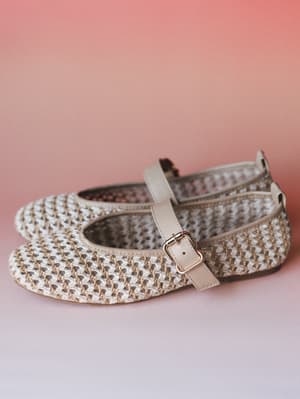 A pair of woven raffia Mary Jane flats in natural cream and tan sit against a soft pink gradient backdrop — the kind of shoes that make you want to plan a picnic immediately.