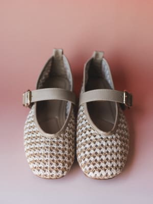 A pair of woven Mary Jane flats in natural cream and tan shot from above against a soft dusty rose background — the kind of shoe that makes an outfit feel intentional without trying too hard.