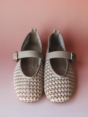A pair of woven Mary Jane flats in natural cream and tan shot from above against a soft dusty rose background — the kind of shoe that makes an outfit feel intentional without trying too hard.
