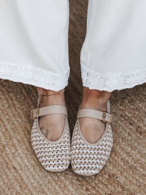 A close-up shot from above of woven Mary Jane flats in natural tan paired with white wide-leg pants trimmed in floral embroidered lace, shot on a jute rug — the whole thing feels like a slow Sunday morning with good intentions.