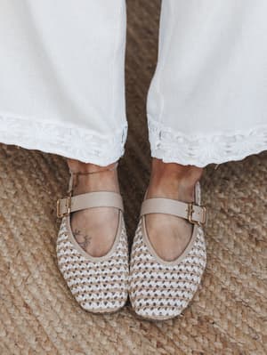 A close-up shot from above of woven Mary Jane flats in natural tan paired with white wide-leg pants trimmed in floral embroidered lace, shot on a jute rug — the whole thing feels like a slow Sunday morning with good intentions.
