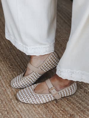A close-up of woven raffia-style Mary Jane flats with a gold buckle strap, paired with wide-leg white pants finished in delicate floral embroidery trim — shot on a jute rug for an effortlessly earthy, collected feel.