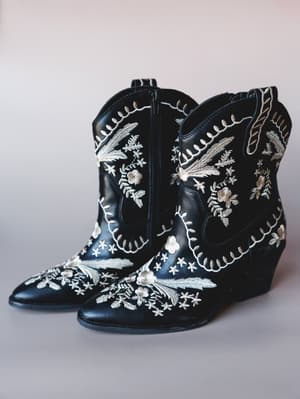 A pair of black leather ankle cowboy booties covered in cream and taupe floral embroidery, shot clean against a soft grey-white studio background — the kind of boots that make a midi dress feel like a whole personality.