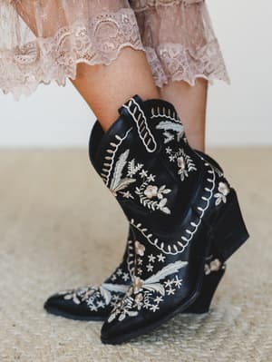 Black western-style booties covered in cream and blush floral embroidery, paired with a dusty pink lace-hem skirt — the kind of detail shot that makes you want to look down at your own feet.