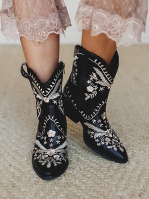 Black leather cowboy booties covered in cream and blush floral embroidery, paired with the hem of a dusty pink lace dress — the kind of boots that do half the outfit's emotional heavy lifting.