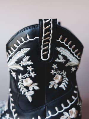 A close-up detail shot of a black leather cowboy bootie covered in intricate cream and tan floral embroidery — the kind of boot that makes a whole outfit without even trying.