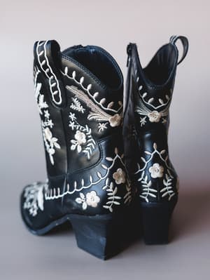 A pair of black leather ankle cowboy booties covered in delicate cream and taupe floral embroidery — the kind of boots that make a plain outfit feel like a whole personality.