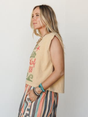 A side-profile shot of a model in a faded mustard graphic muscle tank tucked loosely over multicolor striped wide-leg pants, styled with stacked boho bracelets and a gold chain necklace — easy, sun-warmed, and effortlessly put-together.