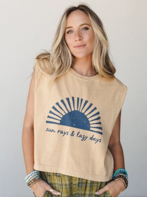A cropped muscle tank in faded mustard with a distressed navy sun graphic and 'sun rays & lazy days' script pairs effortlessly with plaid olive bottoms and a stack of beaded bracelets — the kind of outfit that looks this good without trying.