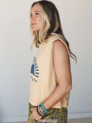 A side-profile shot of a model in a faded mustard cropped muscle tank with a navy sun graphic, styled with olive plaid bottoms and a stack of beaded bracelets — relaxed, warm, and effortlessly lived-in.
