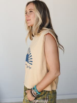 A side-profile shot of a model in a faded mustard cropped muscle tank with a navy sun graphic, styled with olive plaid bottoms and a stack of beaded bracelets — relaxed, warm, and effortlessly lived-in.
