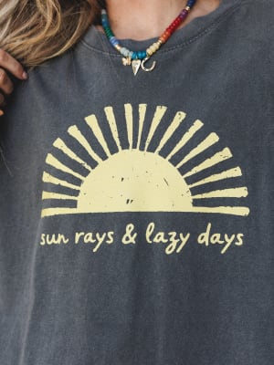 A close-up of a washed charcoal graphic sweatshirt reading 'sun rays & lazy days' with a faded sun illustration, styled with a colorful beaded charm necklace — the kind of top that makes a Tuesday feel a little more intentional.