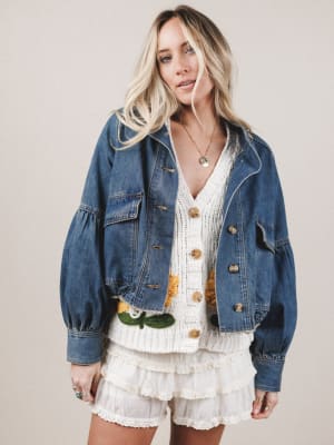 Woman wearing a denim jacket over a white outfit with floral details on a plain background