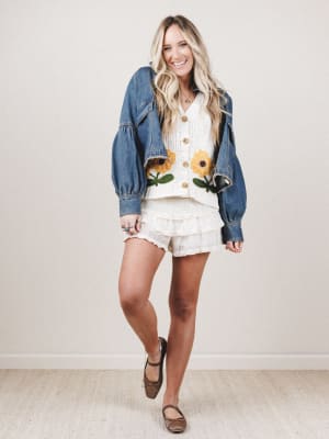 Woman wearing a denim jacket over a white sweater with sunflower designs, standing against a plain background.