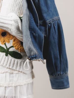 Blue denim jacket draped over a white surface with a textured white garment and floral-patterned object in the background.