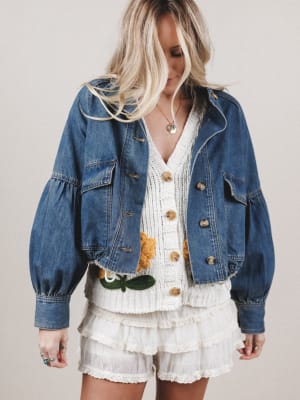 Woman wearing a denim jacket over a white dress with floral details on a plain background