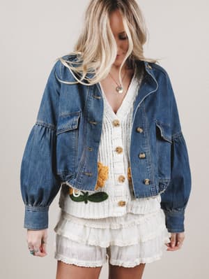 Woman wearing a denim jacket over a white dress with floral details on a plain background