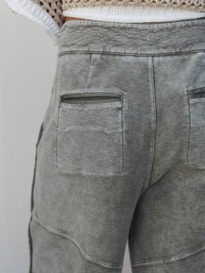 A close-up back detail shot of washed olive barrel pants with patch pockets and zipper closures, paired with a white crochet top — the kind of pants that look like they've been lived in, in the best possible way.