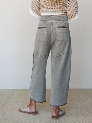 Shot from the back, a model wears wide-leg barrel pants in a washed olive with visible seam detailing and patch pockets, paired with an open-knit cream top and sandals — relaxed and intentional, like a weekend that has nowhere to be.
