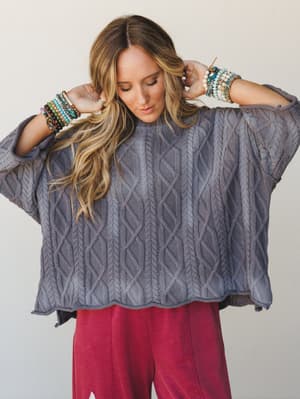 An oversized cable-knit sweater in dusty slate blue-grey worn with raspberry wide-leg pants, styled with armfuls of stacked beaded bracelets — the kind of cozy-but-put-together outfit that feels intentional without trying too hard.