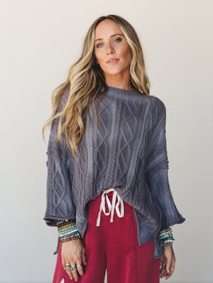 A cable-knit oversized sweater in washed slate grey styled with raspberry-red drawstring pants and stacked beaded bracelets — the kind of effortless, cozy outfit that somehow looks intentional.