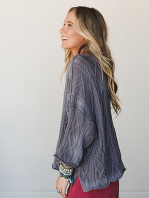 A laughing model shot in profile wearing a slouchy cable-knit sweater in dusty slate blue-grey, layered over a deep magenta bottom, accessorized with a stacked mix of turquoise and stone bracelets — the kind of outfit that feels like a really good Sa