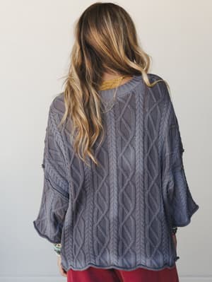 Shot from behind, this dusty slate cable-knit sweater has an oversized, relaxed drape with flared sleeves and a scalloped hem — layered over a mustard top with red pants and stacked turquoise bracelets for an effortlessly expressive, cozy-meets-colle