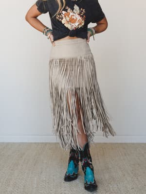 A full fringe suede midi skirt in soft dove tan styled with a knotted floral graphic tee, embroidered cowboy boots, and stacked turquoise bracelets — the whole outfit reads like a music festival dream you actually want to wear to dinner.
