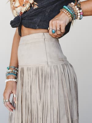 A close-up torso shot of a fringed suede midi skirt in dove grey, styled with a knotted graphic crop top and layered with an abundance of beaded bracelets and turquoise stone rings — the kind of outfit that feels as intentional as it does effortless.